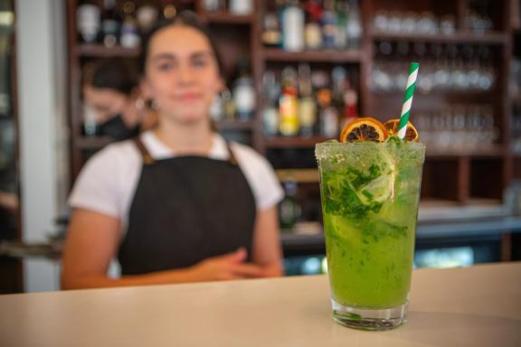 A green cocktail sits towards the camera with a waiter blurred out in the background, and Long beach.
