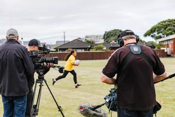 Behind the scenes on the set of Wellington-based reality TV show 'Extreme Cake Sports'. Three crew members stand in the foreground, operating camera and audio equipment while a contestant in a yellow T-shirt runs through the field holding a rugby ball.