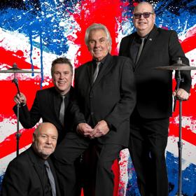 The four members of the UK rock band, Herman’s Hermits, pose between two drum cymbals in front of a Union Jack with a paint splatter effect in the background.