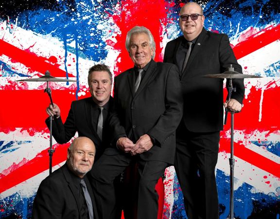 The four members of the UK rock band, Herman’s Hermits, pose between two drum cymbals in front of a Union Jack with a paint splatter effect in the background.