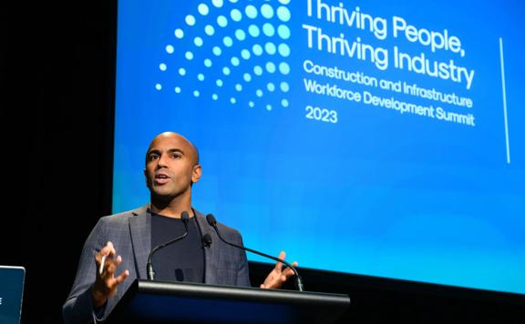 Jehan Casinader, keynote speaker at a business conference stand behind a podium. The projector screen behind him reads 'Thriving People, Thriving Industry Construction and Infrastructure  Workforce Development Summit.