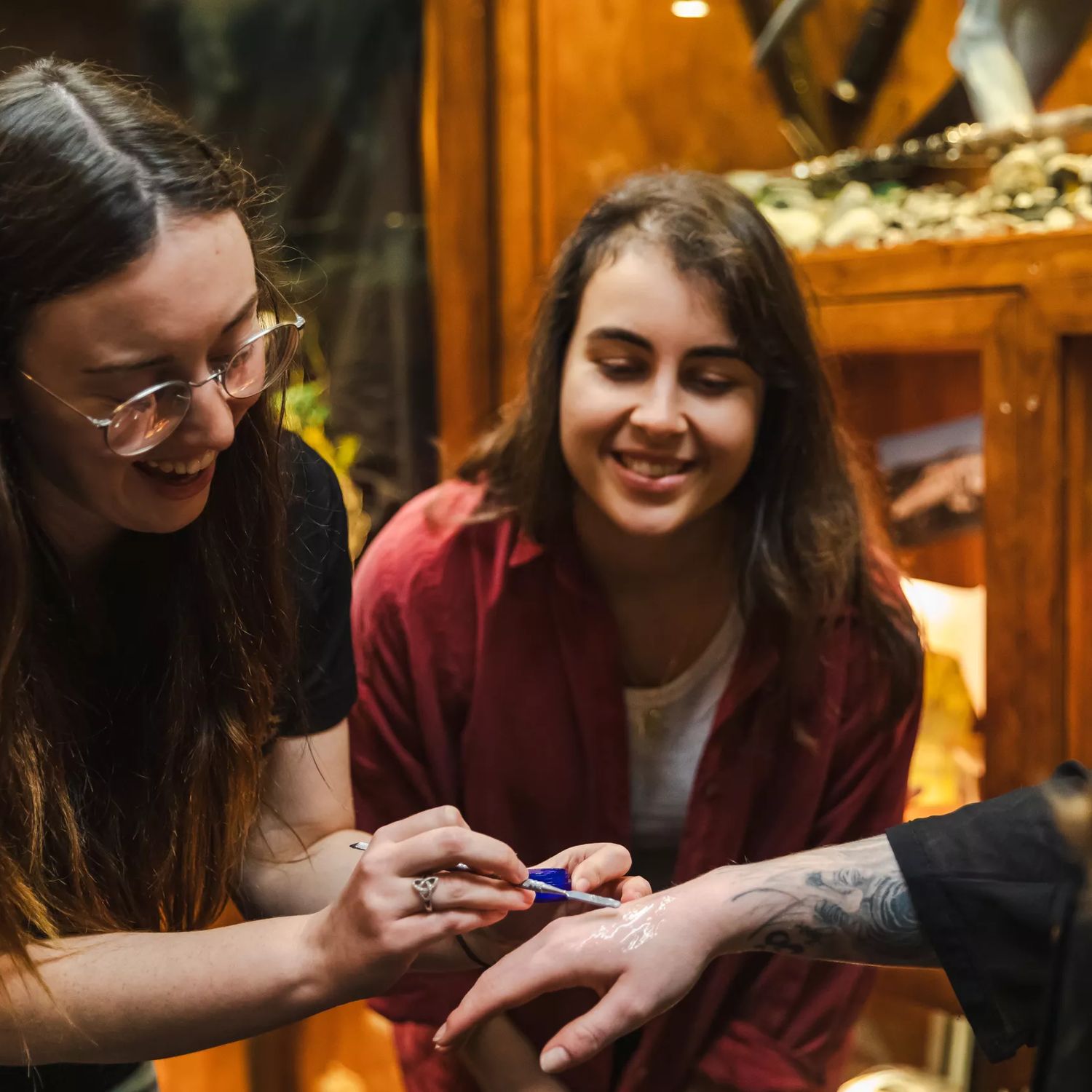 A special effects makeup artist applies a product to someone's hand with a smiling onlooker standing by.