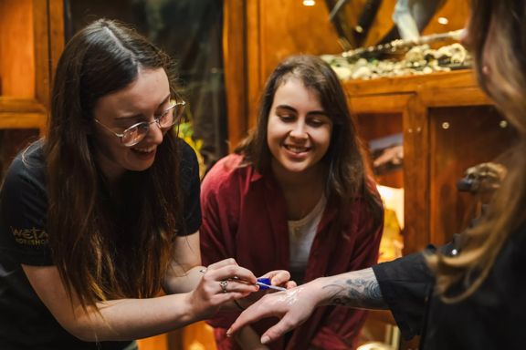 A special effects makeup artist applies a product to someone's hand with a smiling onlooker standing by.