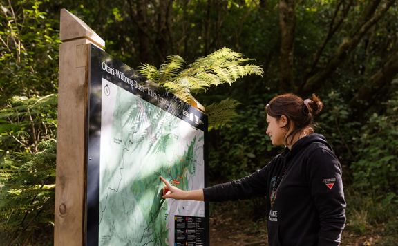 A section of trail Te Ara o Tama in Ōtari-Wiltons Bush. A person is pointing at the path on a green and white sign.
