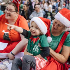 A young child sits on a person's lap, both are wearing Santa hats and enjoying a Christmas performance.