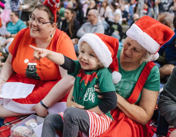 A young child sits on a person's lap, both are wearing Santa hats and enjoying a Christmas performance.