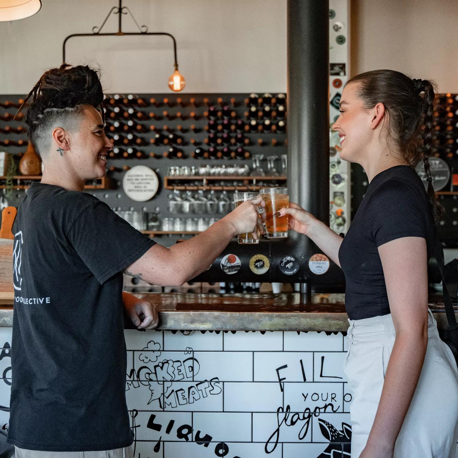 Two people clink glasses at North End Brewery with a wall of bottles filling the back wall behind them.