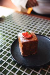 A square pastry with sliced strawberry on top sits on a black plate on a green tile table.