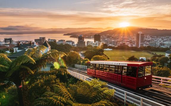 Wide of the cable car driving up the tracks with the sun setting over Wellington city and harbour in the background.