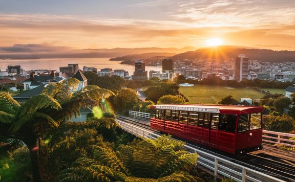 Wide of the cable car driving up the tracks with the sun setting over Wellington city and harbour in the background.