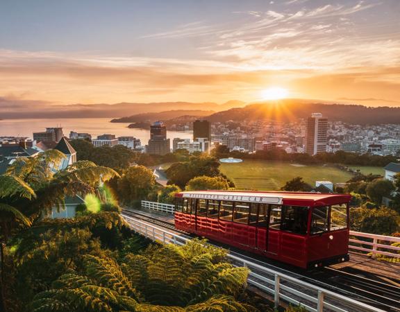 Wide of the cable car driving up the tracks with the sun setting over Wellington city and harbour in the background.