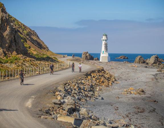 Cyclists are riding along a coastal road at the Pencarrow Coast in Eastbourne, New Zealand.