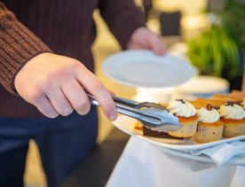 A person wearing blue pants and a brown jumper, is using tongs to pick up a cupcake and put it onto a small white plate at the James Cook Hotel Grand Chancellor.