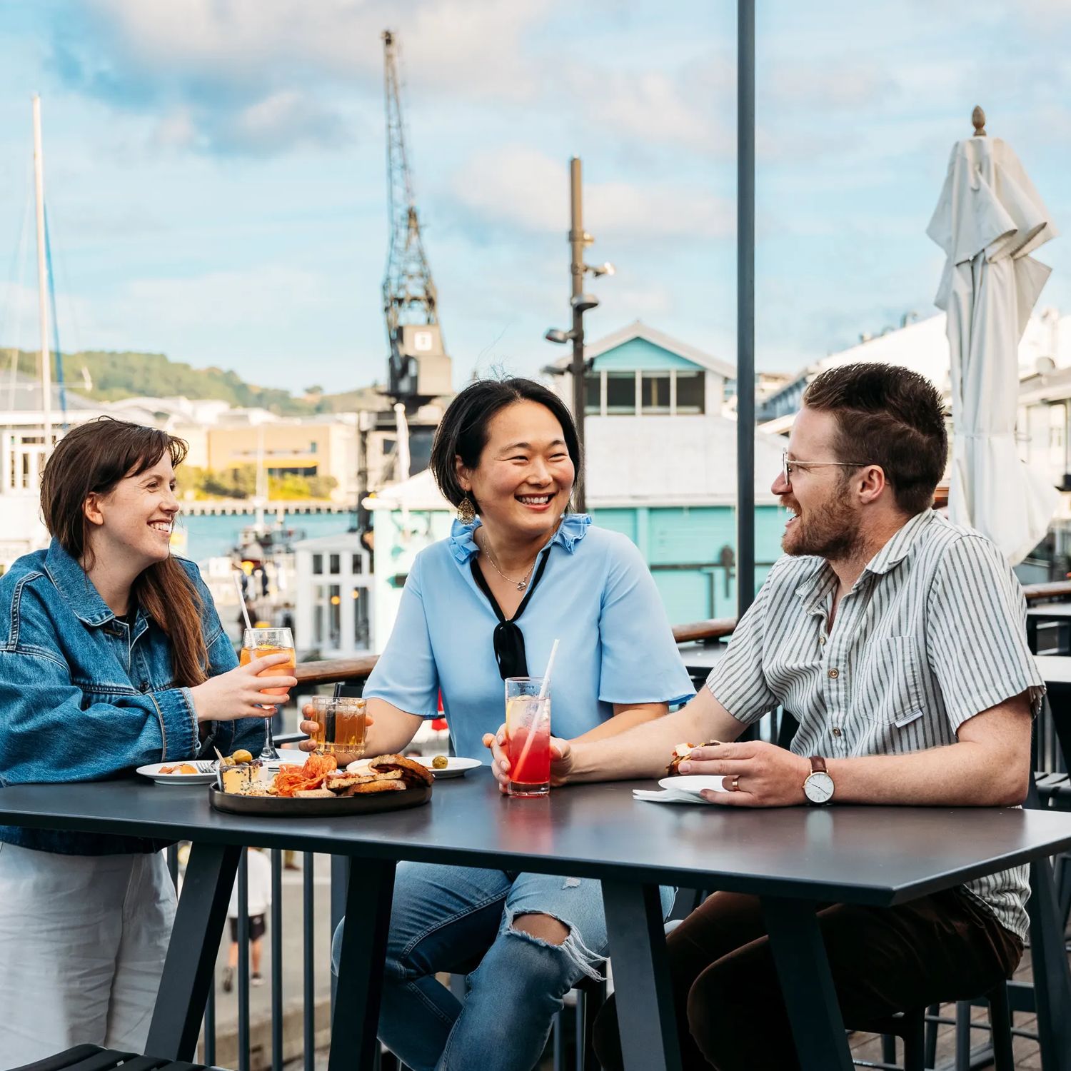 Three people sit at a bar table at Foxglove Bar on the Wellington Waterfront. They are all drinking a cocktail and a charcuterie board sits between them.