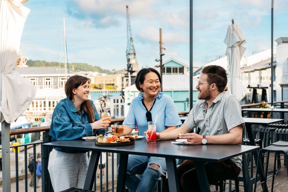 Three people sit at a bar table at Foxglove Bar on the Wellington Waterfront. They are all drinking a cocktail and a charcuterie board sits between them.