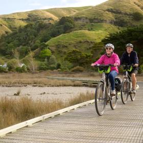 Two people riding bikes on Te Ara Piko Walkway. The trail features stretches of boardwalk and bridges, with amazing views of the harbour. 