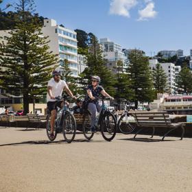 Two people ride bicycles along Oriental Bay Parade in Wellington on a sunny day.