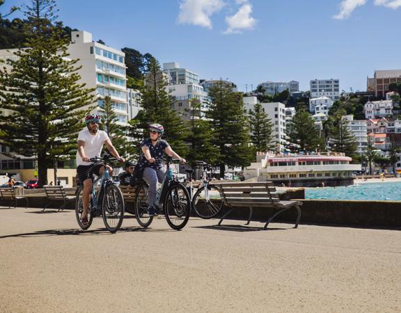 Two people ride bicycles along Oriental Bay Parade in Wellington on a sunny day.