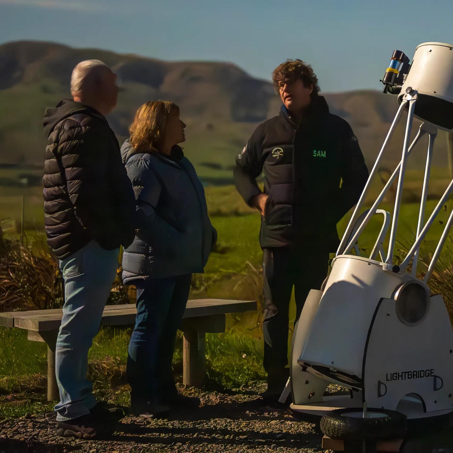 Sam Leske and two guests at Star Safari stand and chat by a telescope in an open Wairarapa valley.