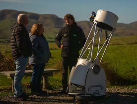 Sam Leske and two guests at Star Safari stand and chat by a telescope in an open Wairarapa valley.