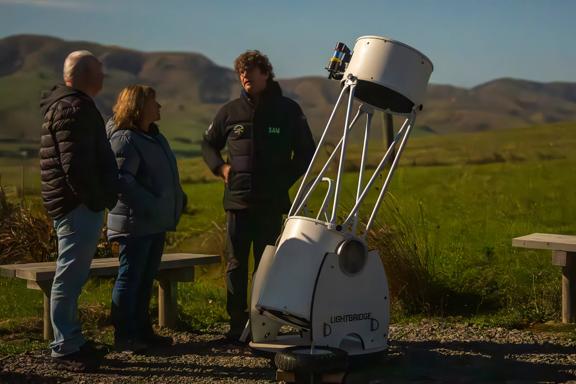 Sam Leske and two guests at Star Safari stand and chat by a telescope in an open Wairarapa valley.