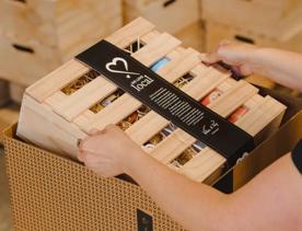 A person who is out of frame places a wooden box with a black label wrapped around it into a larger cardboard box with more wooden boxes arranged into piles in the background.