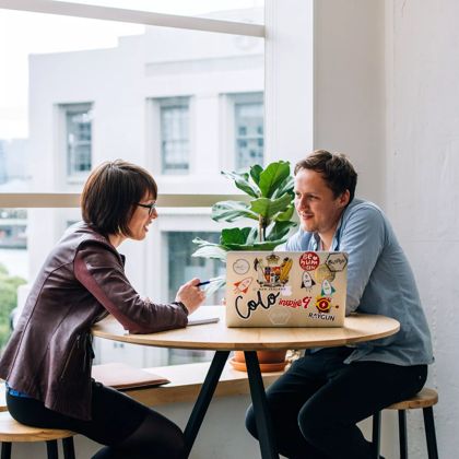 Two people sit and chat at a high table, with a laptop beside a large window.