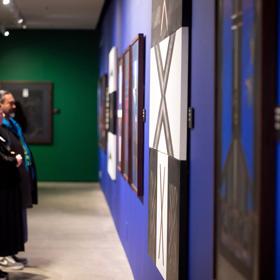 An exhibition at the Pātaka Art + Museum located in Porirua City Centre featuring large painted canvases displayed on the walls. Four people stand and admire the artwork.