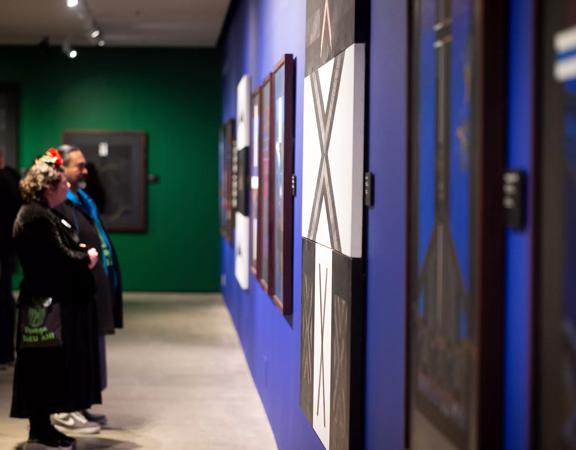 An exhibition at the Pātaka Art + Museum located in Porirua City Centre featuring large painted canvases displayed on the walls. Four people stand and admire the artwork.