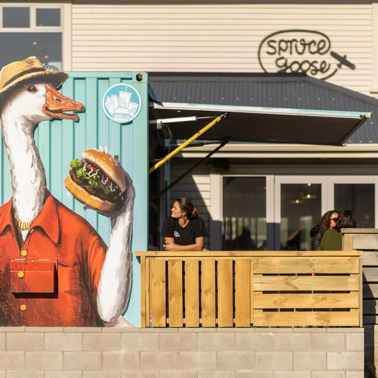 A person stands with their elbows on the fence of Spruce Goose's patio looking at a mural of a Goode wearing a fedora, holding a surfboard in one hand and a burger in the other. 