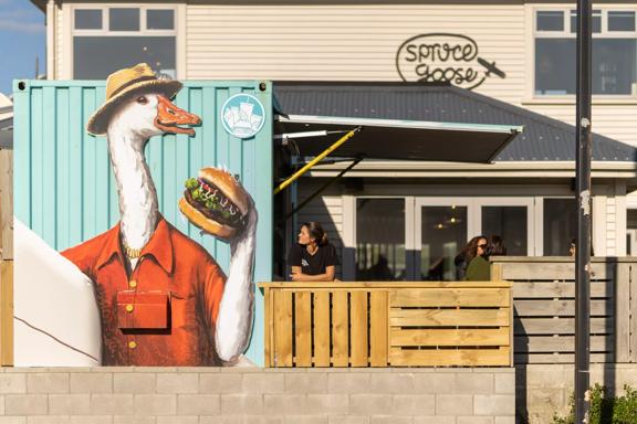 A person stands with their elbows on the fence of Spruce Goose's patio looking at a mural of a Goode wearing a fedora, holding a surfboard in one hand and a burger in the other. 