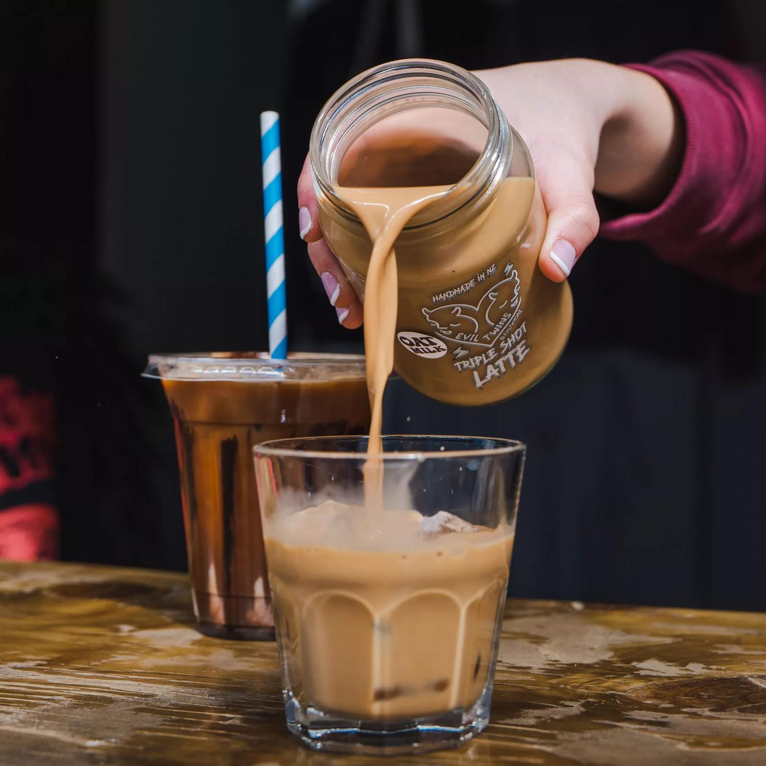 An iced coffee being poured from a glass jar, with another iced coffee sitting nearby in a plastic cup, made by Evil Twins.