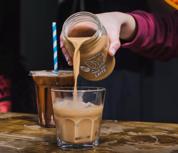 An iced coffee being poured from a glass jar, with another iced coffee sitting nearby in a plastic cup, made by Evil Twins.