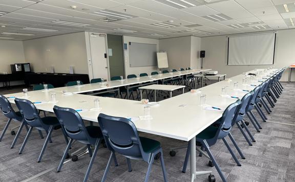 A big conference room set up with tables in a U shape, chairs, notepads, pens, and water glasses. 