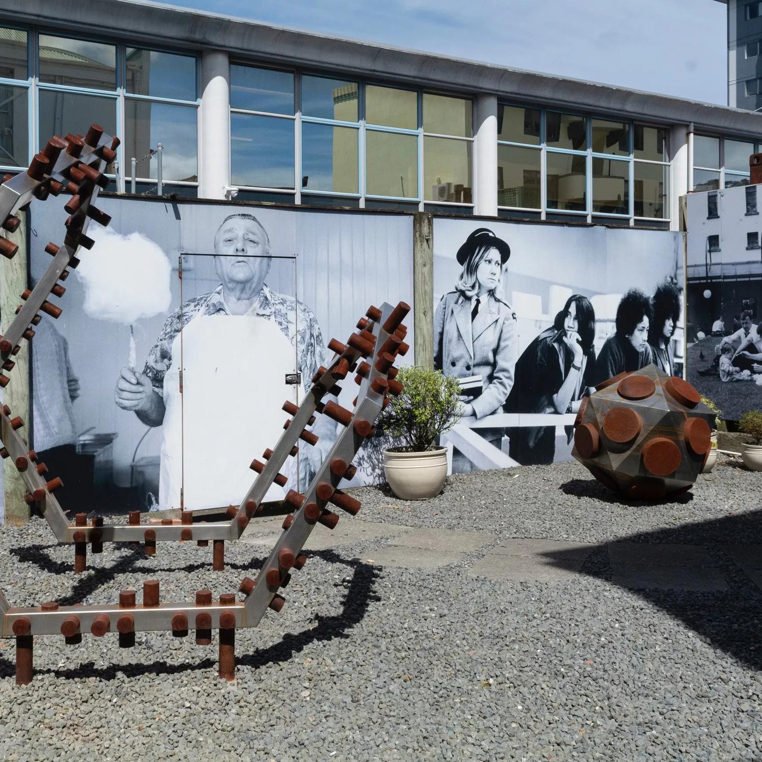 Image of the sculpture yard behind Suite Gallery. The walls are  covered in huge black and white photographic images. The ground is covered in gravel and has two large corten steel sculptures.