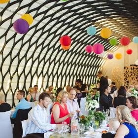 People are seated at two long tables with white tablecloths in the Kamala function room at Wellington Zoo. Colourful lanterns hang from the intricate metal ceiling.