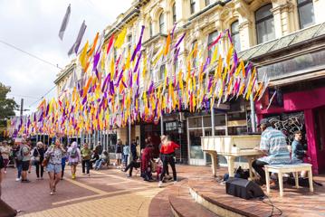 A busker plays a white grand piano for passersby on Cuba Street in Te Aro Wellington with white, yellow, orange, pink and purple streamers suspended from above.