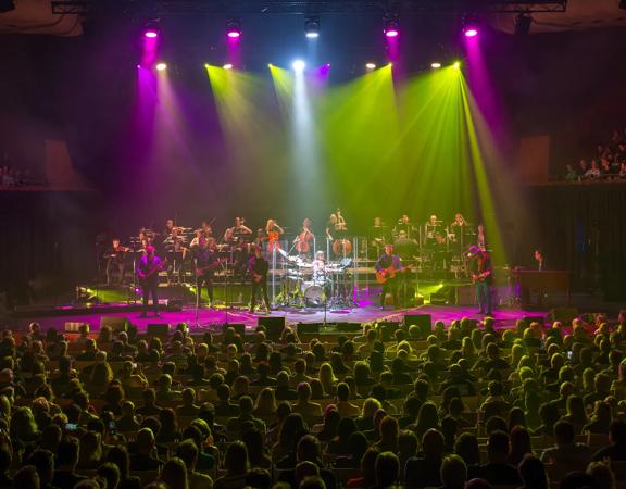 Approximately 20 musicians perform on stage under fuchsia and yellow spotlights to a packed audience.