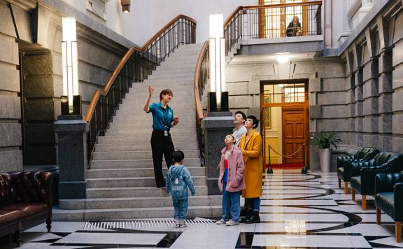 Visitors on a guided tour stand in the entrance hall of New Zealand Parliament in Wellington, with a sweeping staircase and marble floor behind them.