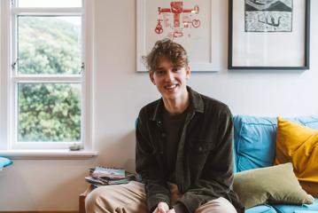 Charlie Faulks, animator of 'Bloke of the Apocolypse' wears a brown tee-shirt, jacket and khaki pants, smiles and sits on a living room couch.