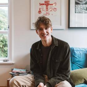 Charlie Faulks, animator of 'Bloke of the Apocolypse' wears a brown tee-shirt, jacket and khaki pants, smiles and sits on a living room couch.