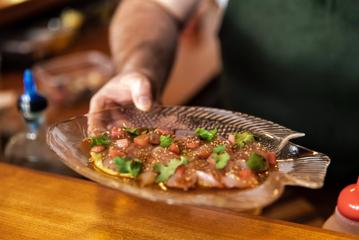 A hand holding a glass fish-shaped dish with a crudo garnished with cilantro.