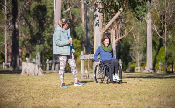 Two people in a flat grassed park area. One is a wheel chair user and the other is standing holding a takeaway coffee cup.