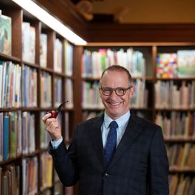 David Sedaris standing in a library between bookshelves, wearing a suit and holding a smoking pipe, with rows of books visible in the background.