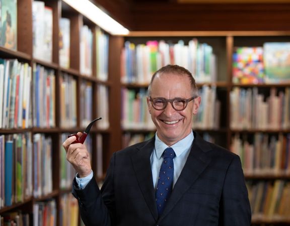 David Sedaris standing in a library between bookshelves, wearing a suit and holding a smoking pipe, with rows of books visible in the background.