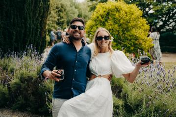 Two people smiling, wearing sunglasses, and holding glasses of rosé wine, pose arm-in-arm.