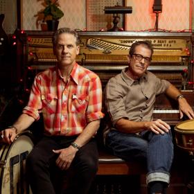 The two main band members of Calexico, an Arizona-based indie rock band, Joey Burns and John Convertino sit on a piano bench in a room surrounded by instruments. 