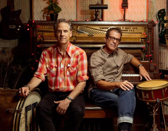 The two main band members of Calexico, an Arizona-based indie rock band, Joey Burns and John Convertino sit on a piano bench in a room surrounded by instruments. 