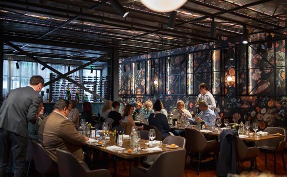 Lunch at La Serre by Sofitel Wellington, people dine on long tables as waitresses cater. A large floral accent wall is seen on the back wall.