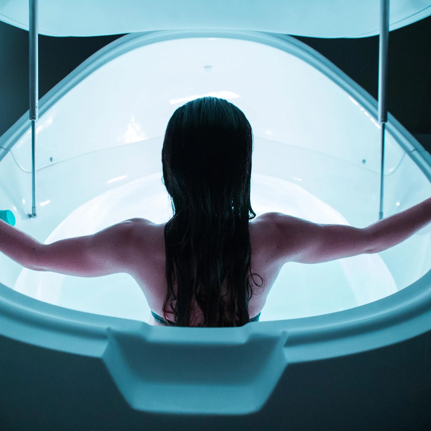 A person in a bathing suit sits facing away inside an illuminated floating tank.